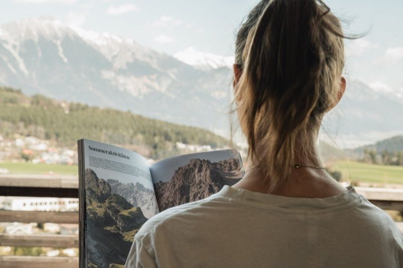 Frau liest auf dem Balkon das Hotelmagazin, im Hintergrund die Berge