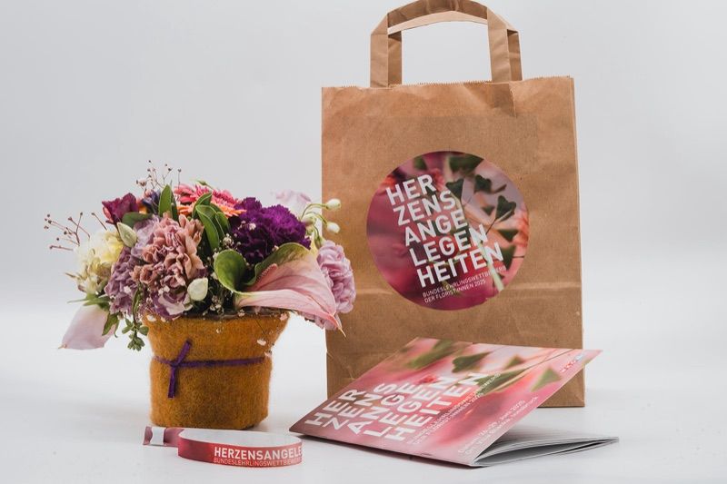 Papiertasche mit Aufkleber „Herzensangelegenheiten“, daneben Blumen, Folder und Armband